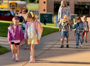 Two young girl students walk hand in hand with back packs on. Two young male students walk behind them with female teacher smiling.